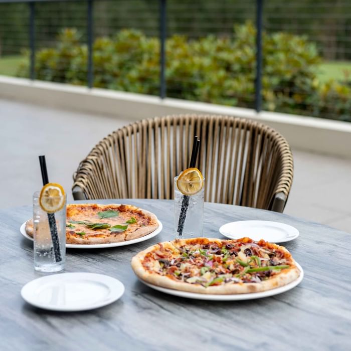 Two pizzas and glasses of water on Karinyas Terrace table with a wicker chair at Mercure Kooindah Waters