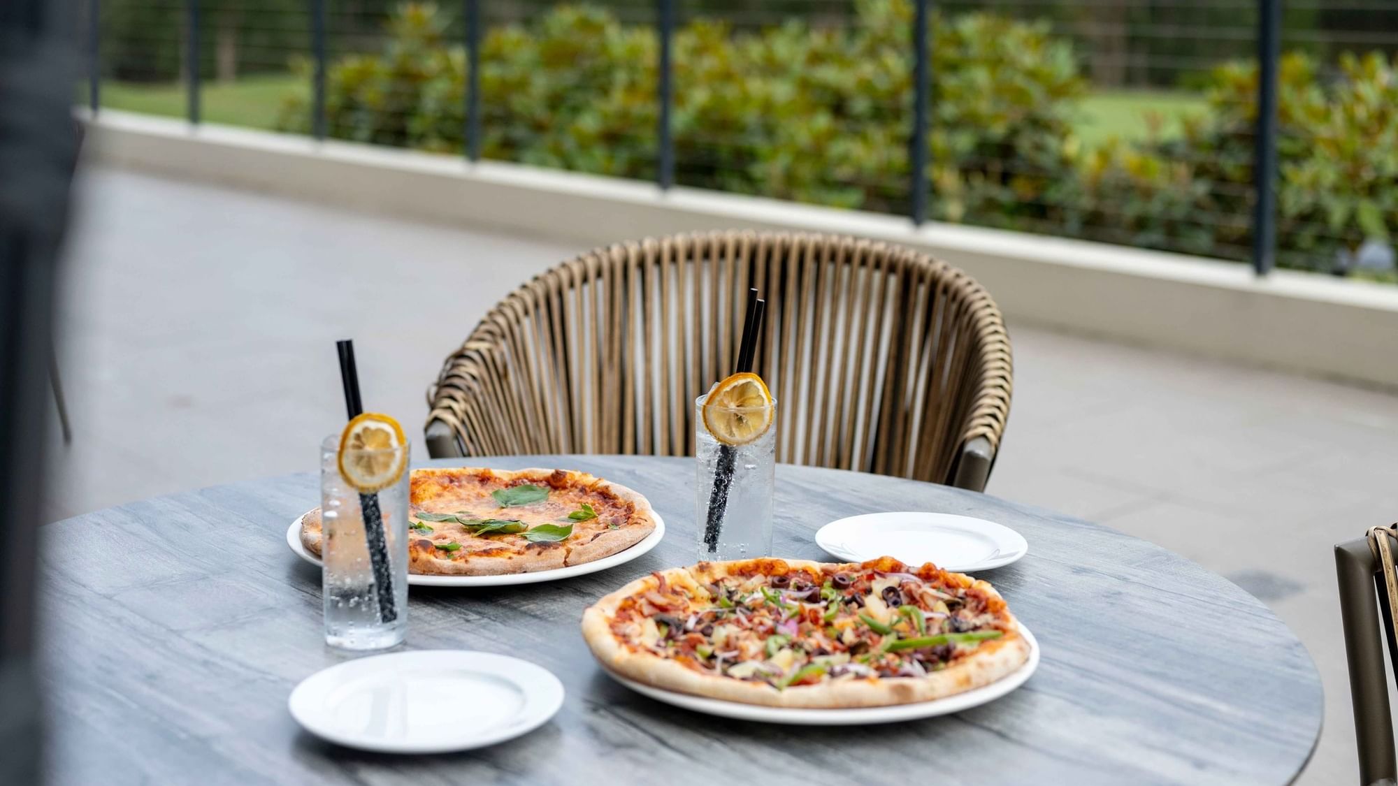 Two pizzas and glasses of water on Karinyas Terrace table with a wicker chair at Mercure Kooindah Waters