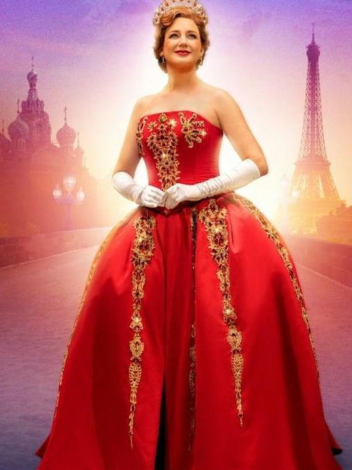 Woman in ornate red ball gown with gold embellishments and crown against backdrop featuring Eiffel Tower and Russian architecture.