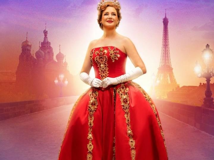 Woman in ornate red ball gown with gold embellishments and crown against backdrop featuring Eiffel Tower and Russian architecture.