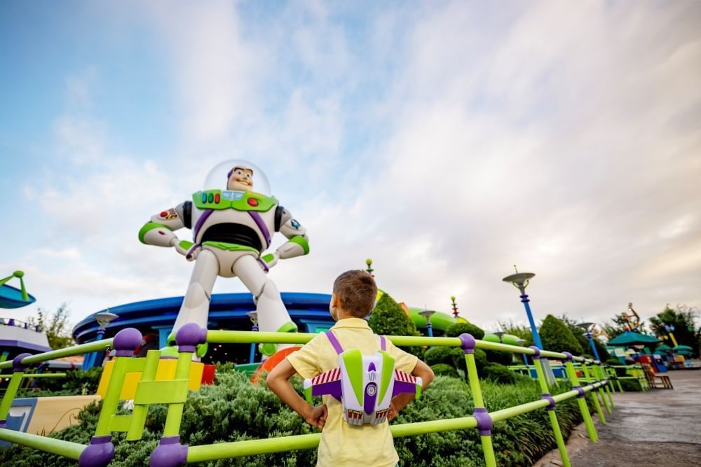 A boy with a Buzz Lightyear backpack stands with his hands on his hips facing a large statue of Buzz Lightyear. 