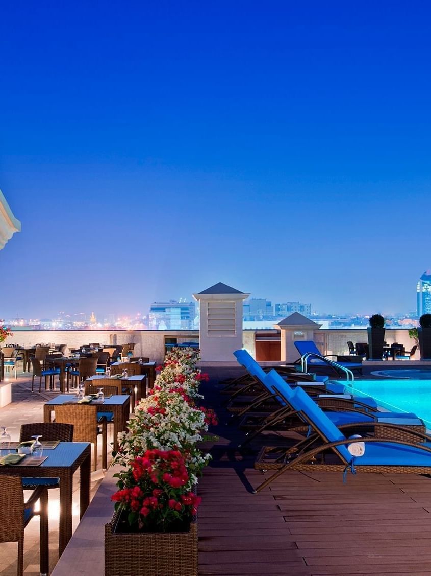 Blue loungers by a glowing pool under a clear night sky surrounding dining tables at Warwick Doha Hotel
