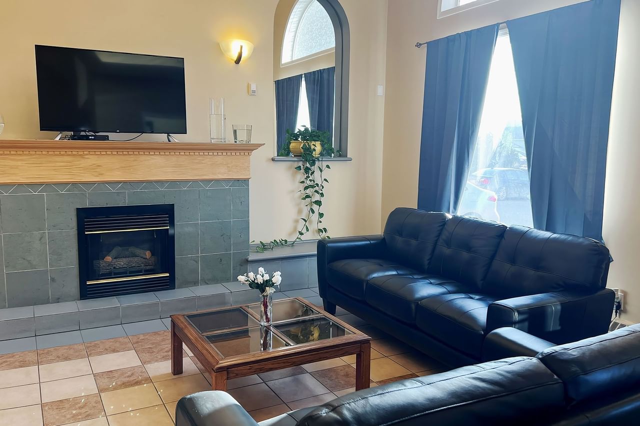 Living room with two black leather couches, glass coffee table, fireplace, TV, and large window with blue curtains.