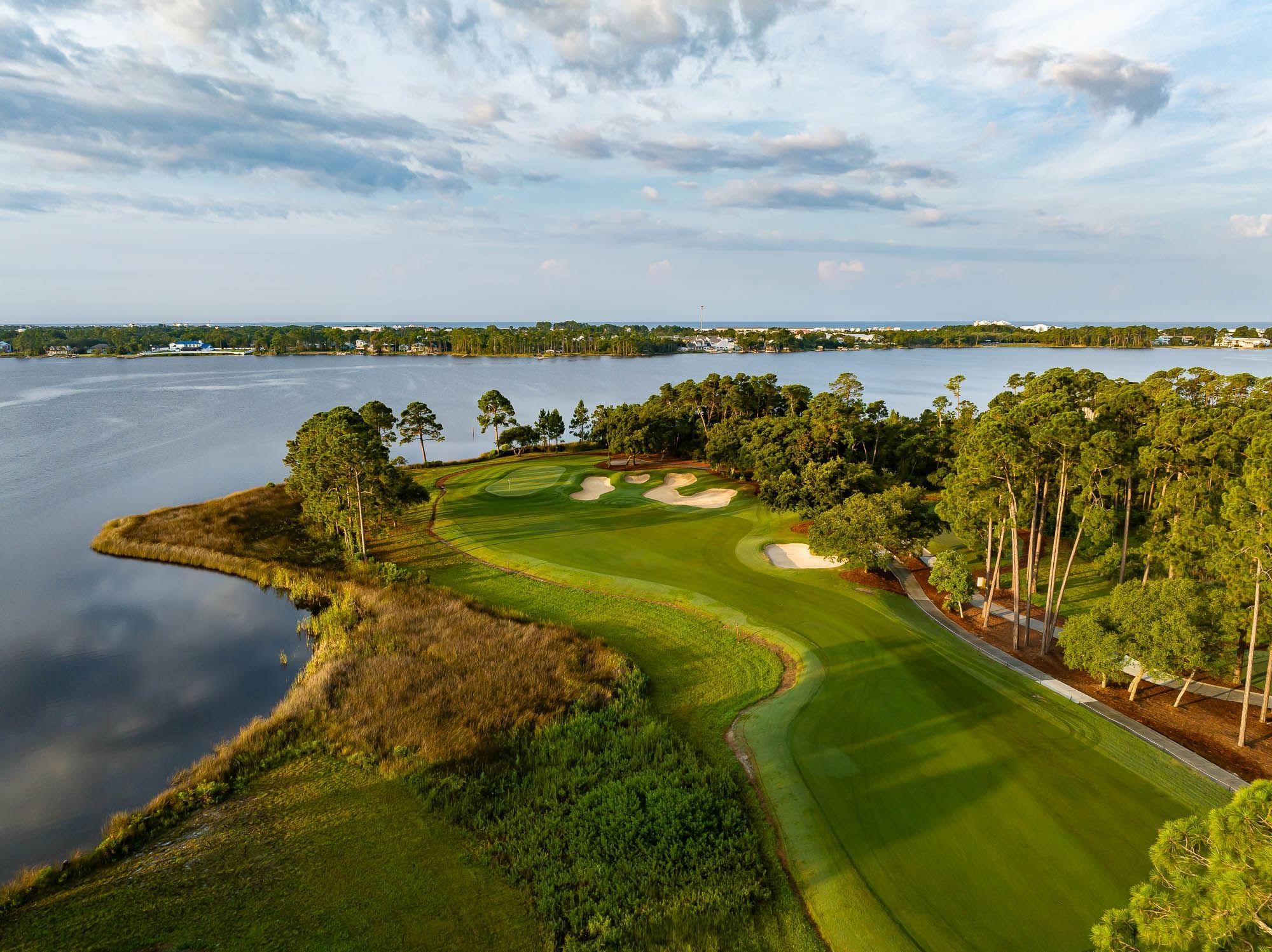 Aerial view of a golf course with water, trees, and a distant city skyline under a cloudy sky.