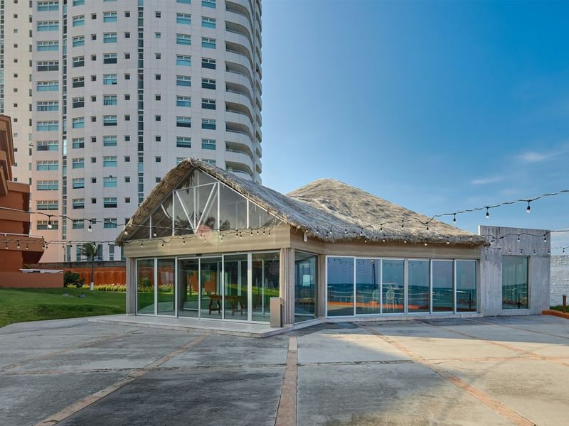 Modern building with thatched roof and large glass walls next to a tall building.