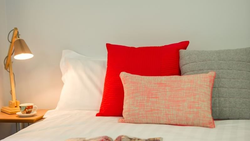 Modern bedroom with white bedding, red accent pillows, wooden desk lamp, and framed wall art at StudentLiving - on Margaret