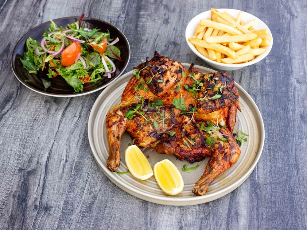 Grilled chicken with lemon wedges, salad, and fries on a wooden surface.