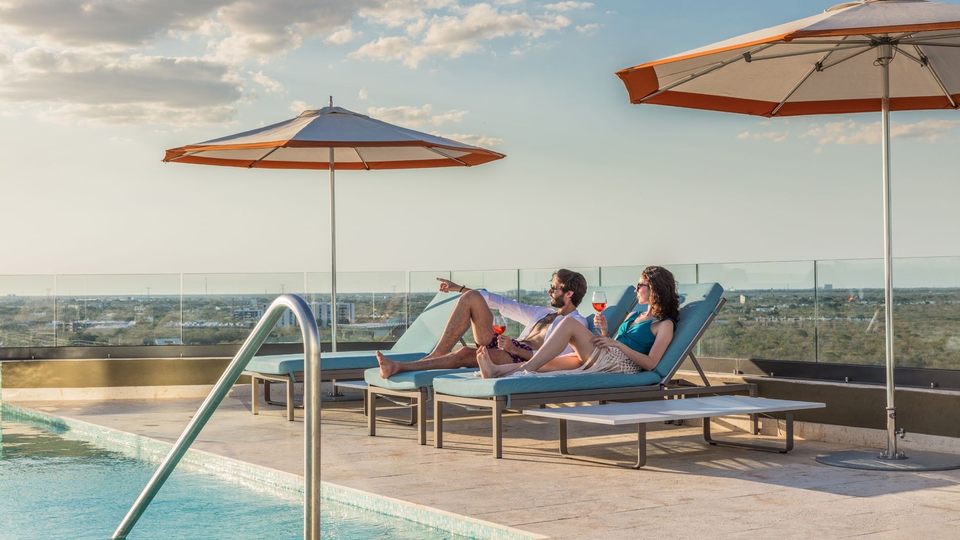 Guests relaxing rooftop pool, lounging on blue chairs under umbrellas with city vistas at Camino Real Merida