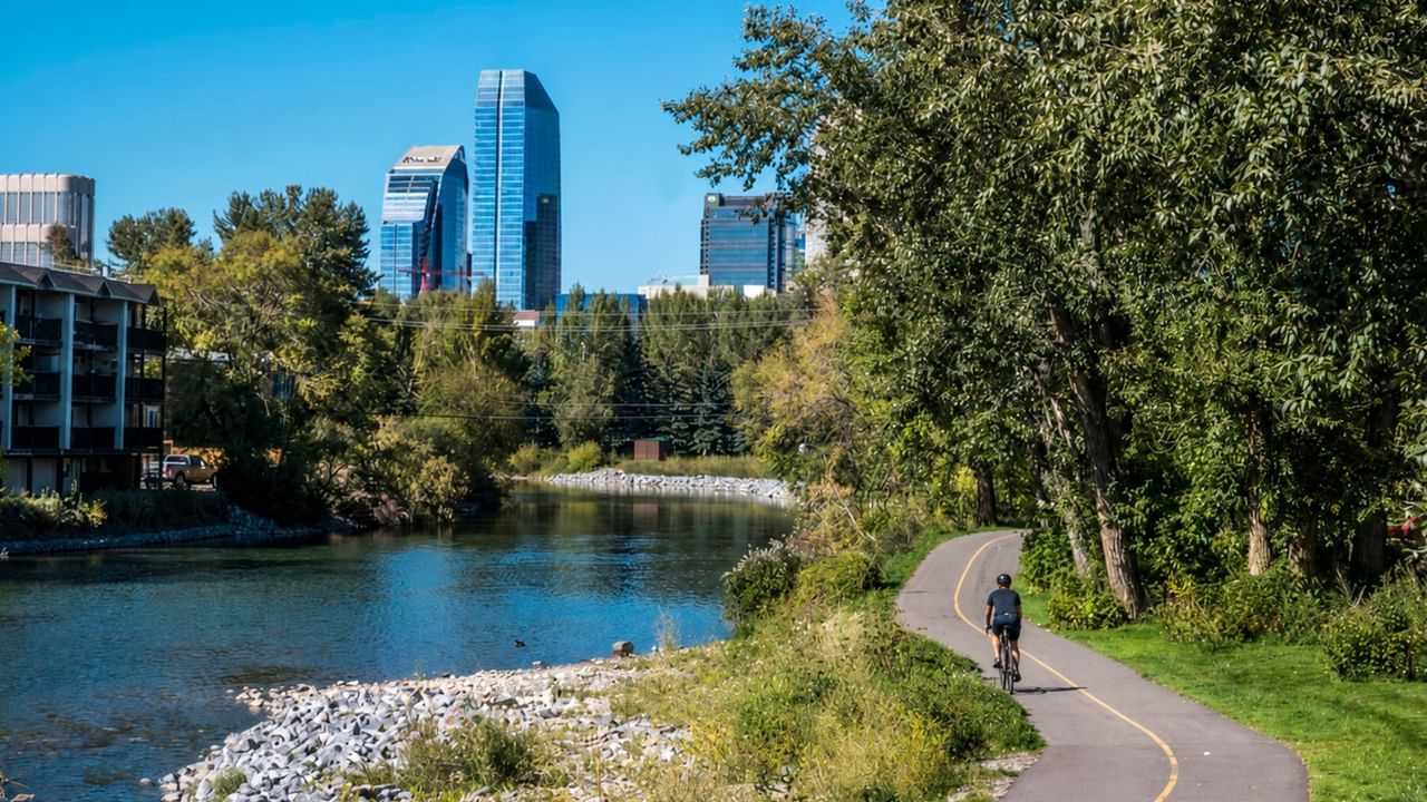 Cyclist riding along Bow River Pathway with city skyline and lush greenery framing the scenic waterfront trail.