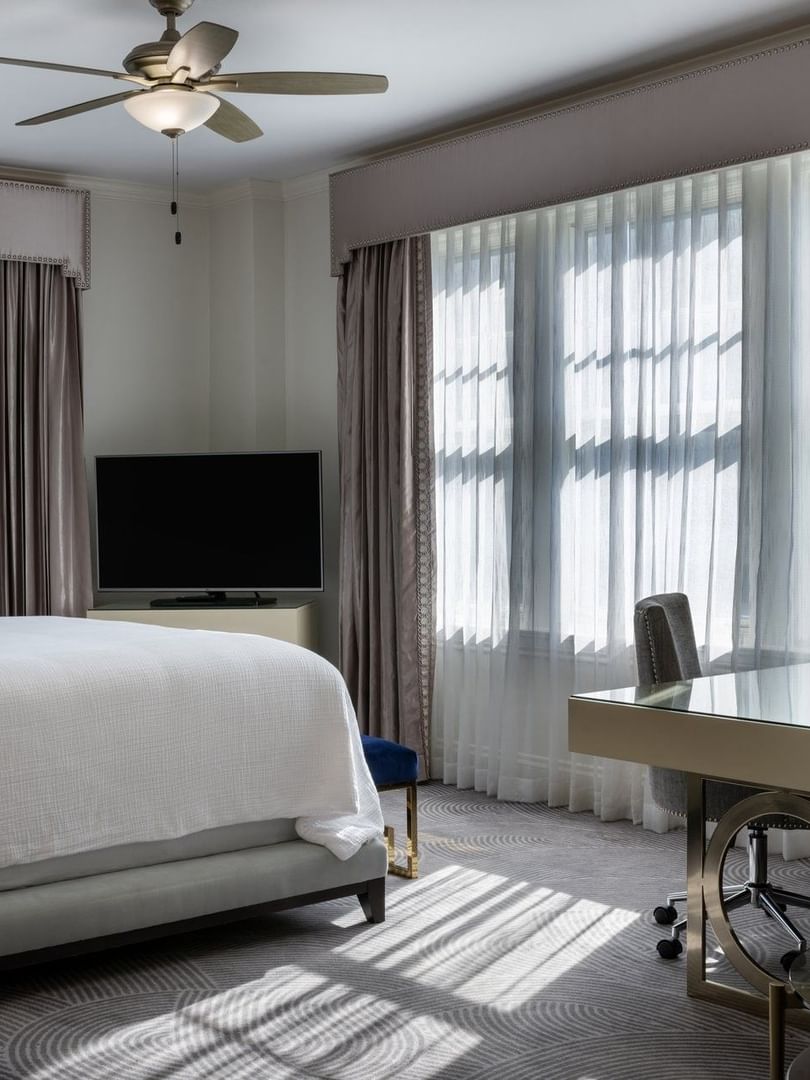 Sleek glass desk by a white bed under a ceiling fan in a bright, modern room in Presidential Suite at Warwick Melrose Dallas