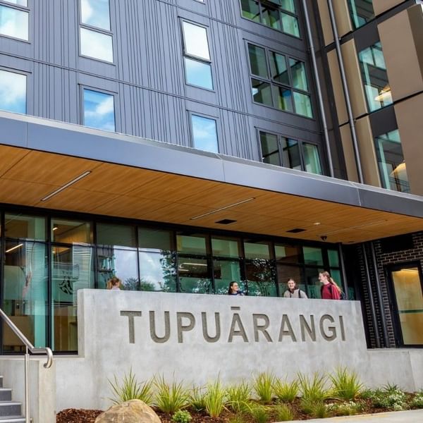 Modern multi-story building entrance with glass doors, wooden canopy, and concrete Tupuārangi sign with guests walking outside.