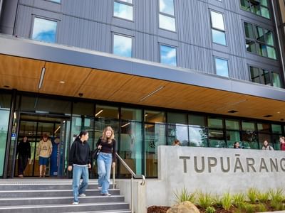 Modern multi-story building entrance with glass doors, wooden canopy, and concrete Tupuārangi sign with guests arriving.