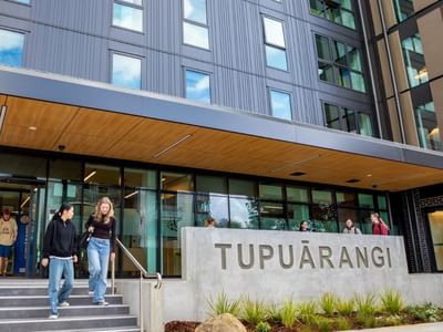 Modern multi-story building entrance with glass doors, wooden canopy, and concrete Tupuārangi sign with guests walking outside.