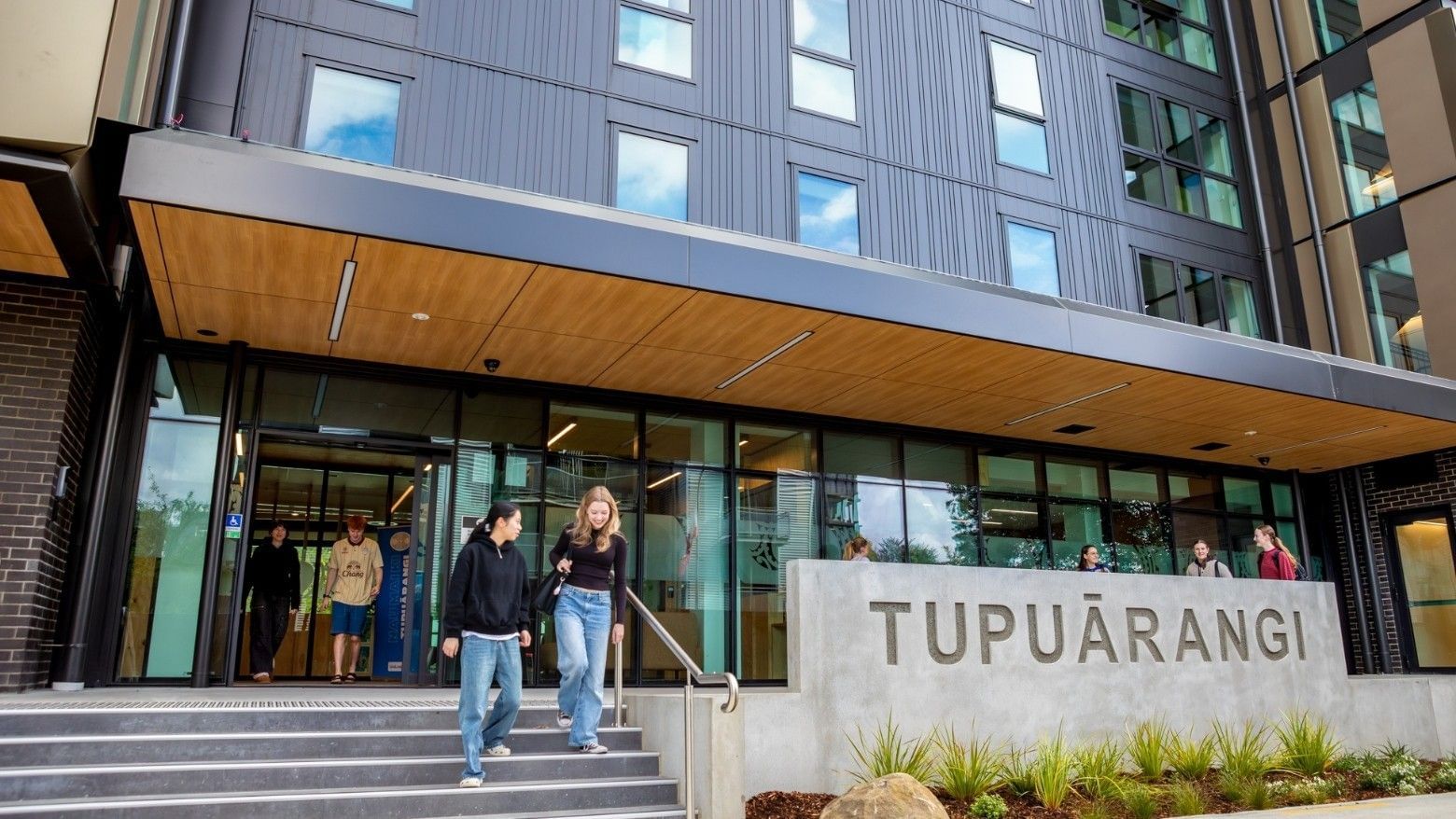 Modern multi-story building entrance with glass doors, wooden canopy, and concrete Tupuārangi sign with guests arriving.