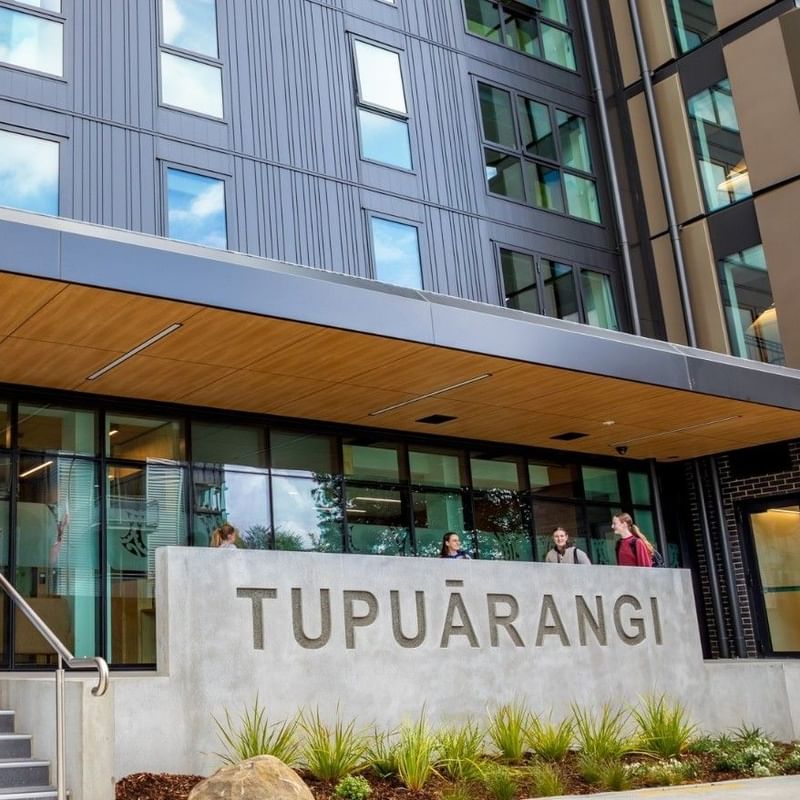 Modern multi-story building entrance with glass doors, wooden canopy, and concrete Tupuārangi sign with guests walking outside.