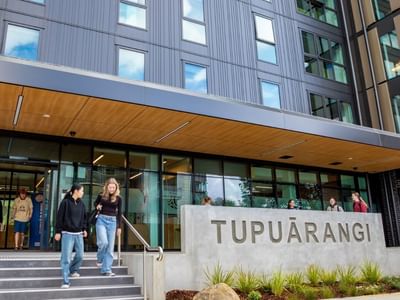 Modern multi-story building entrance with glass doors, wooden canopy, and concrete Tupuārangi sign with guests walking outside.