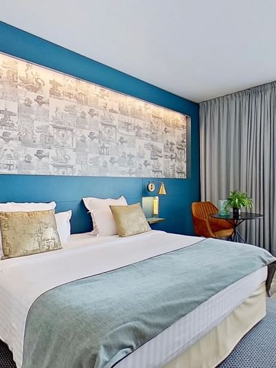 Chambre d'hôtel avec un grand lit, une table, une plante, et une vue sur une fenêtre avec des rideaux.