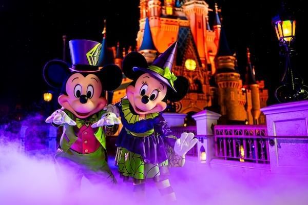 Mickey and Minnie Mouse dressed in Halloween costumes pose in a cloud of purple haze in front of Cinderella's Castle lit with orange lights.