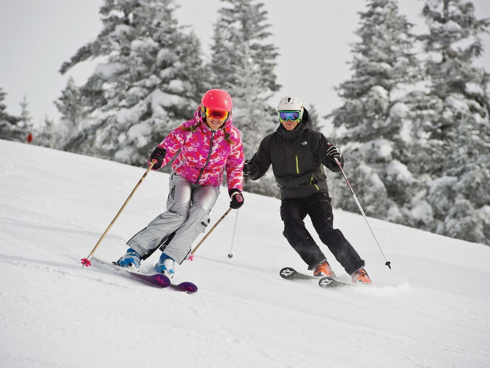 Two skiers enjoying the slopes with snowy pines in the background.