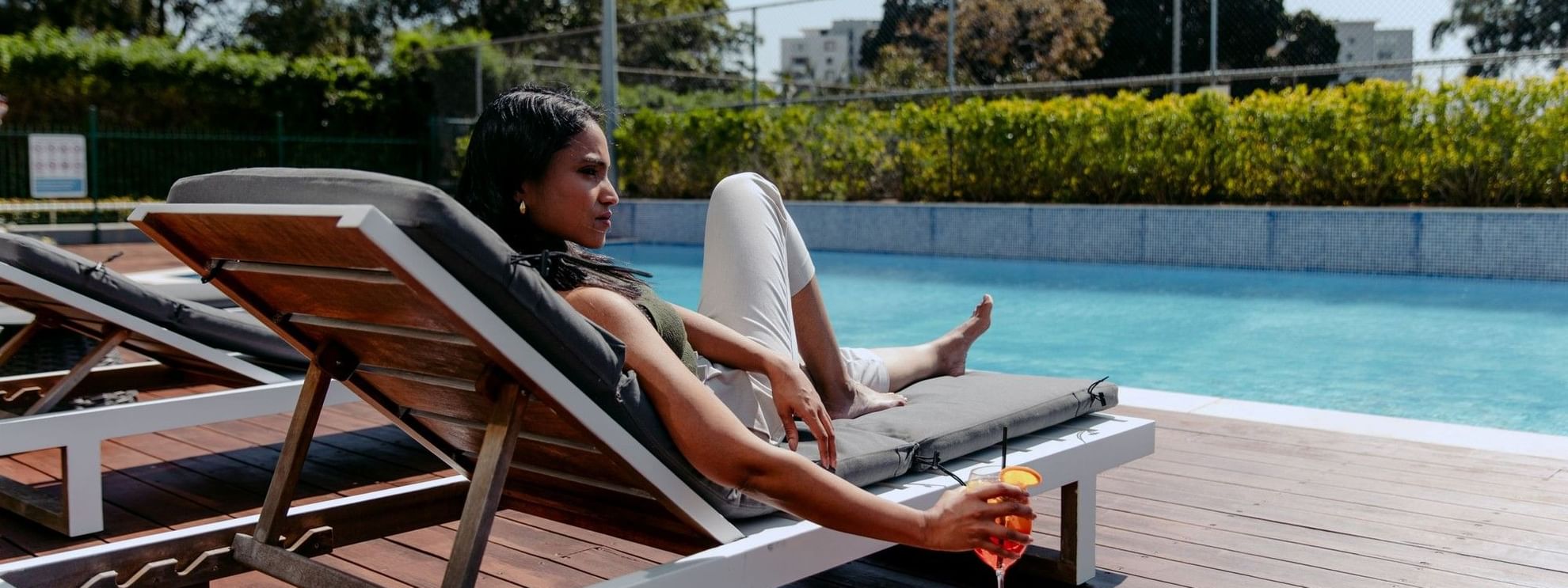Woman lounging on a lounge chair by a pool, holding a cocktail drink.