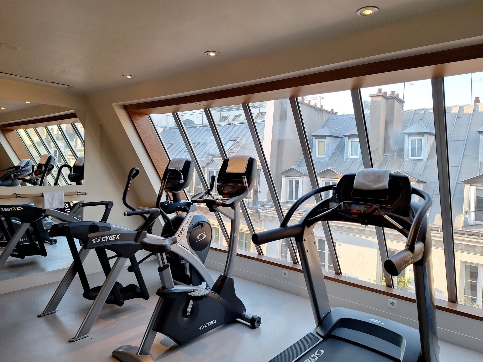 Fitness equipment placed by floor-to-ceiling sloped windows with rooftop views at Hotel Westminster Paris