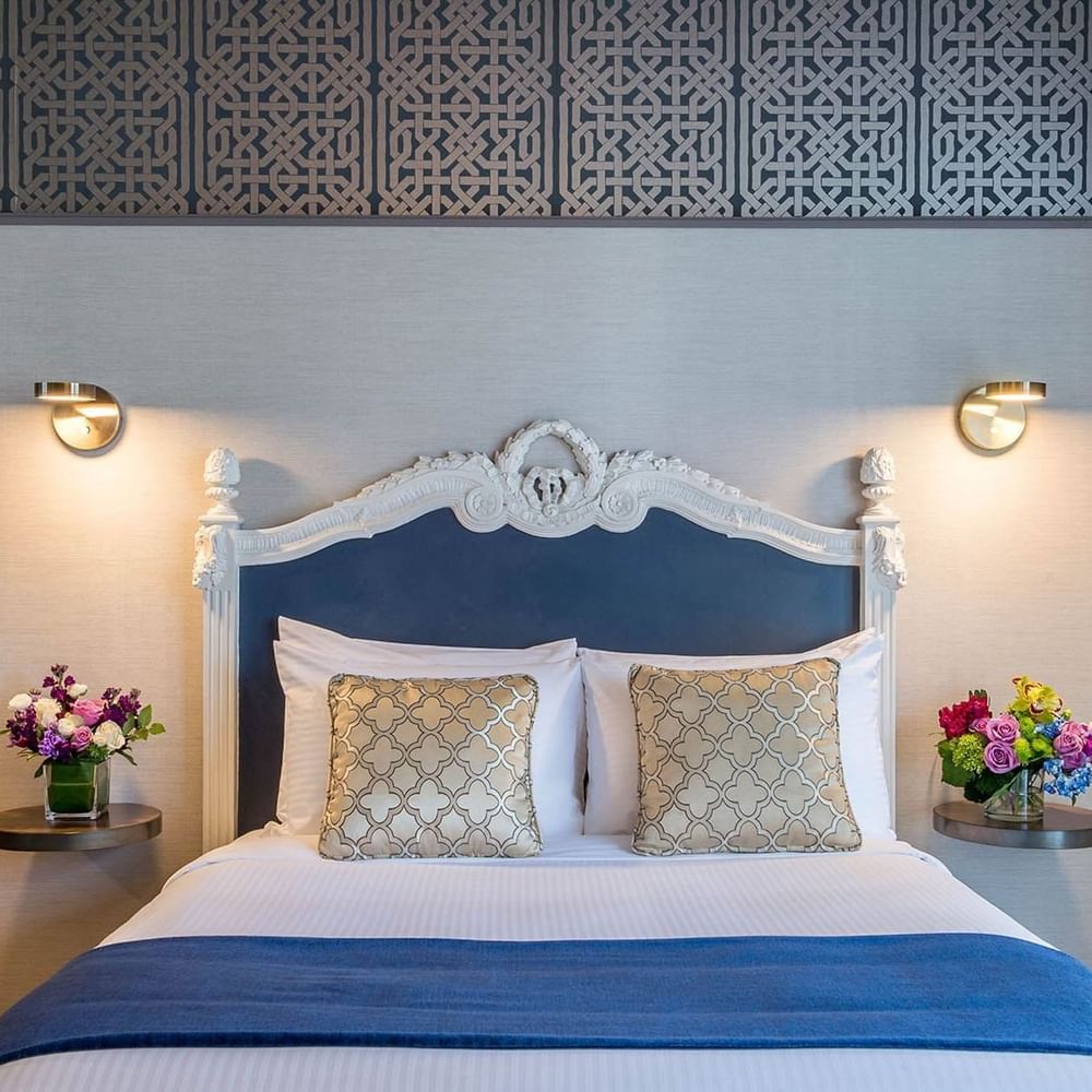 Floral vases on round tables by a bed under an ornate white headboard in Superior Queen Room at Warwick San Francisco
