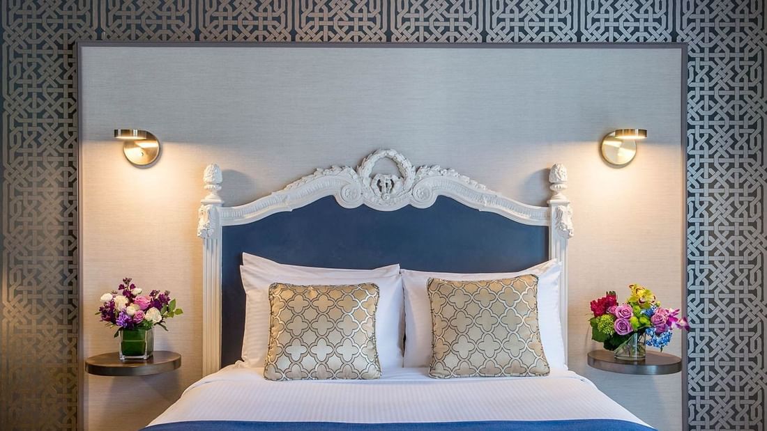 Floral vases on round tables by a bed under an ornate white headboard in Superior Queen Room at Warwick San Francisco