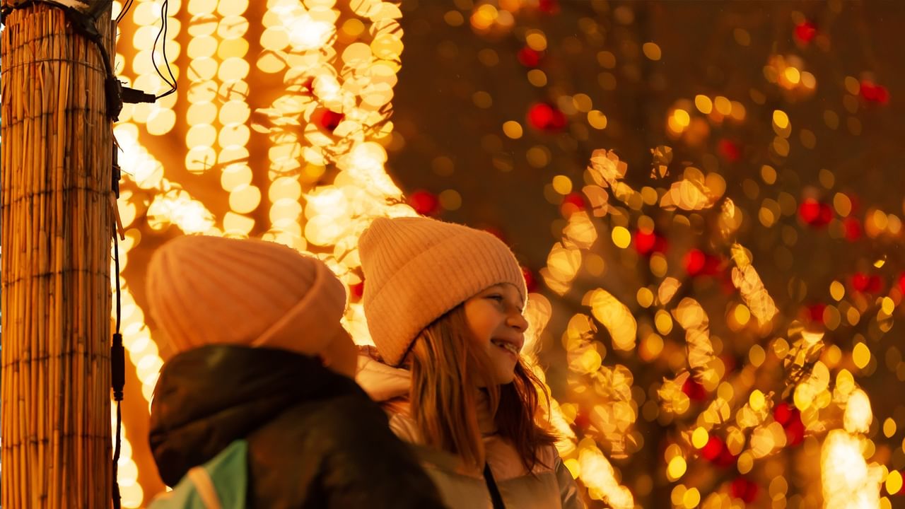 Two children in winter hats stand close together, smiling, in front of a festively lit tree.