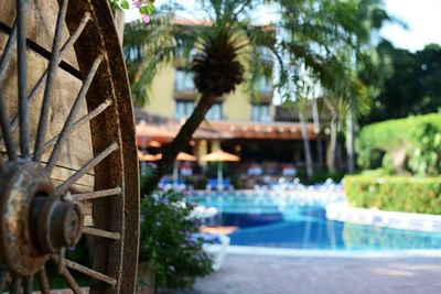 Rusty wagon wheel in front of pool with umbrellas, blue chairs, palm trees, and yellow building in background.