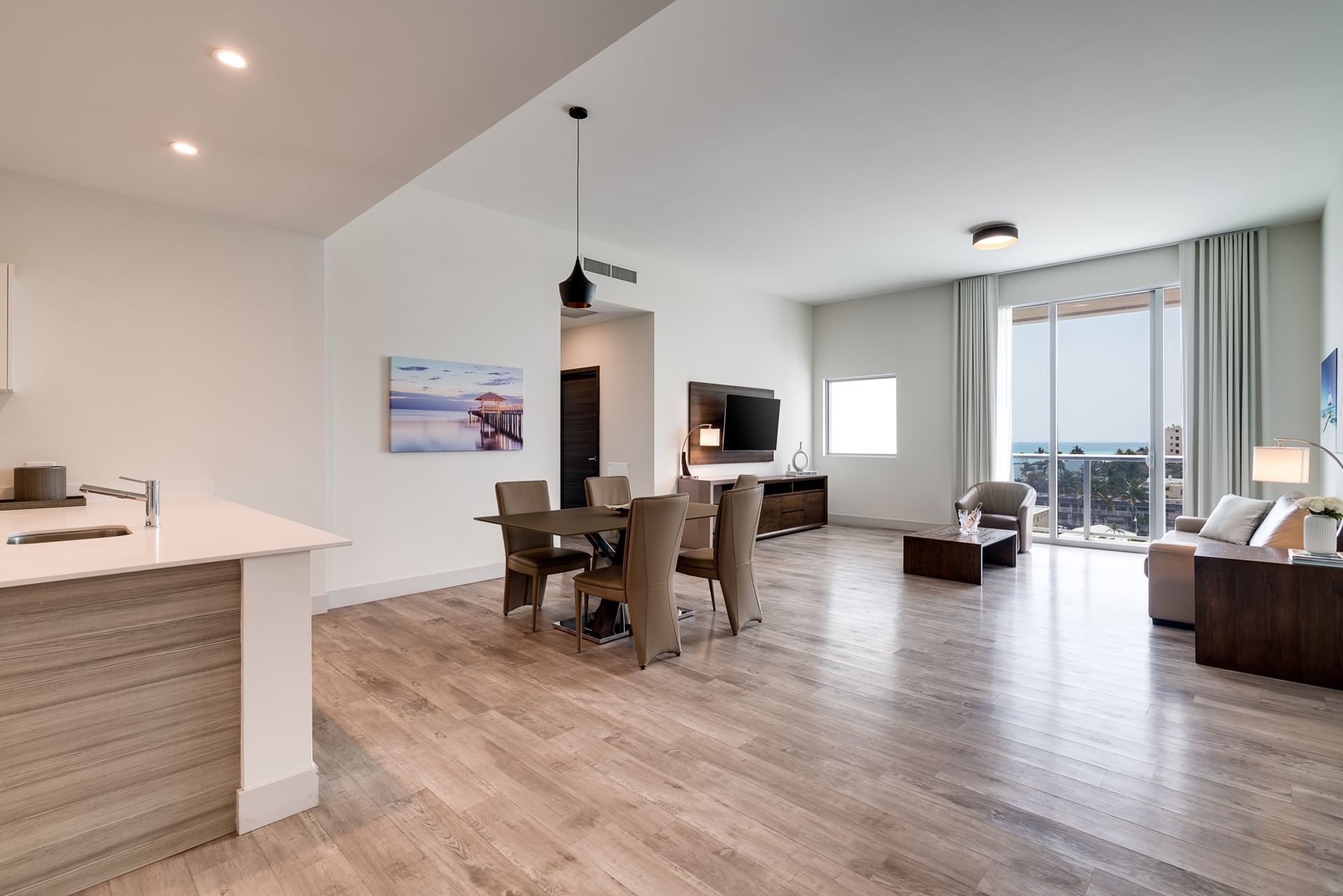 A wide view of a modern living room with kitchenette, dining area, and sliding glass doors leading to a balcony.