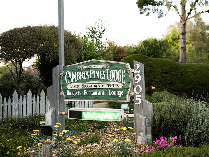 Sign for Cambria Pines Lodge Resort & Conference Center with Vacancy light in front of white picket fence and flowers.