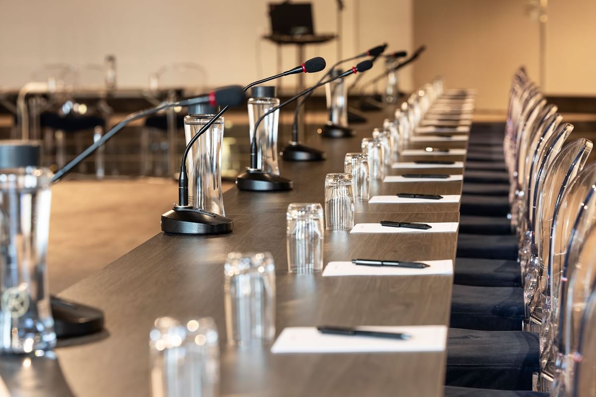 Långt konferensbord med mikrofoner, glas och papper på varje plats, omgärdat av stolar.