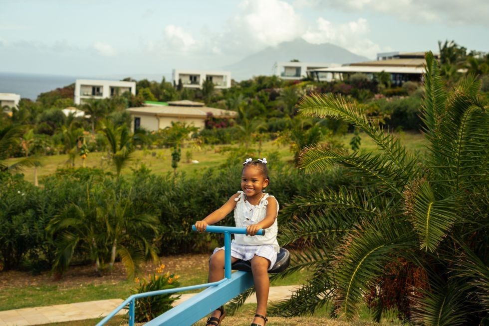 Kind op een blauwe wip in een groen park met palmbomen en villa's op een heuvel nabij Golden Rock Resort