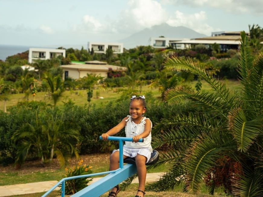 Child on blue seesaw in green park with palm trees and hillside villas near Golden Rock Resort