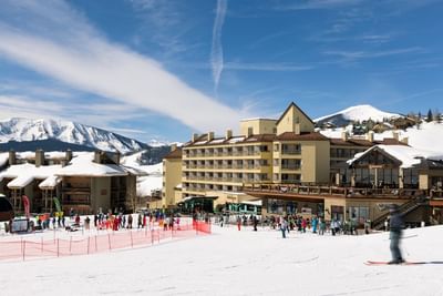 Skier passing resort and crowd of people with snow-covered mountains in background at Elevation Resort & Spa