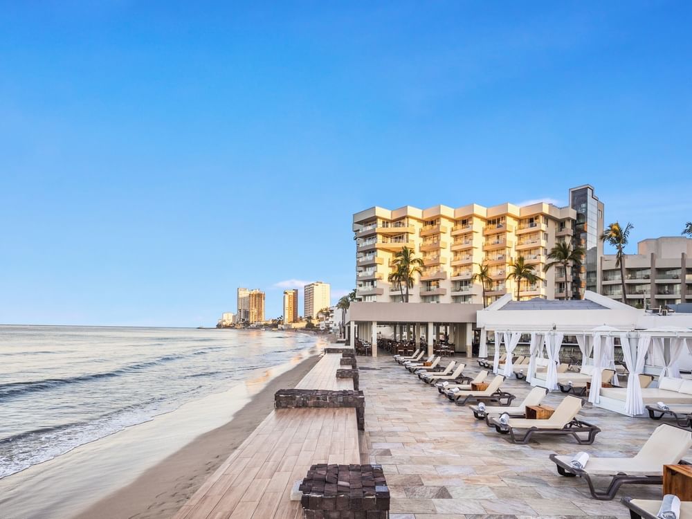 Scenic beach Sunvivia Mazatlán resort view featuring white cabanas & lounge chairs on a stone deck beside the calm shoreline