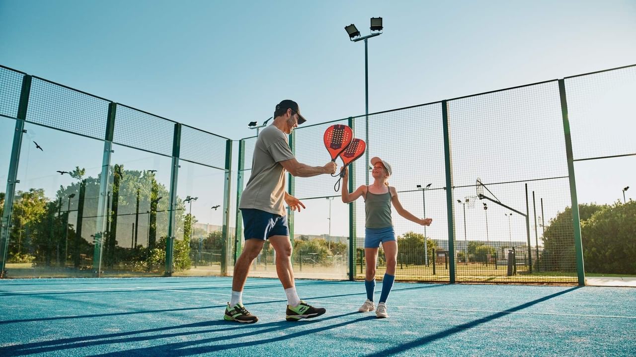 Un uomo e una bambina con racchette da padel su un campo sportivo in una giornata di sole.