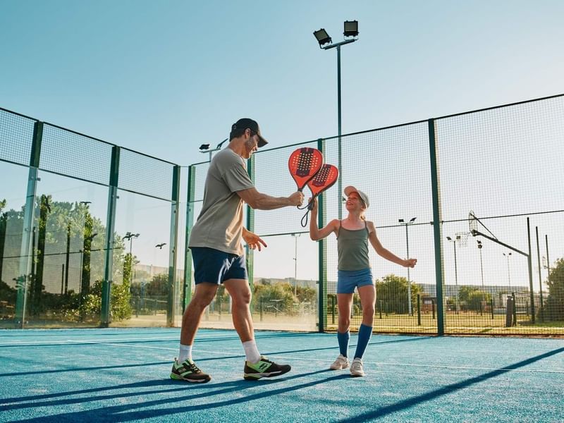 Mann und Mädchen spielen Paddle-Tennis auf einem blauen Platz mit grünen Zäunen.