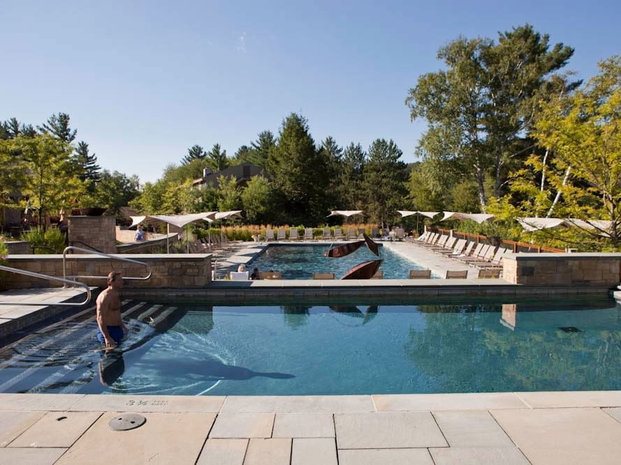 A man stands in a large outdoor pool with lounge chairs, trees, and umbrellas under a clear sky.