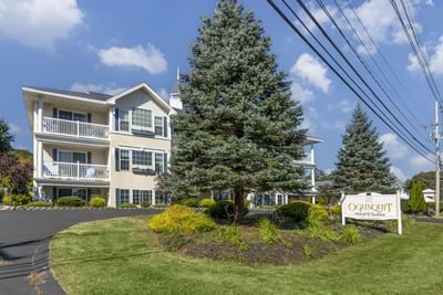 Ogunquit Hotel & Suites exterior with balconies, landscaped grounds, and a sign beneath a blue sky on a sunny day