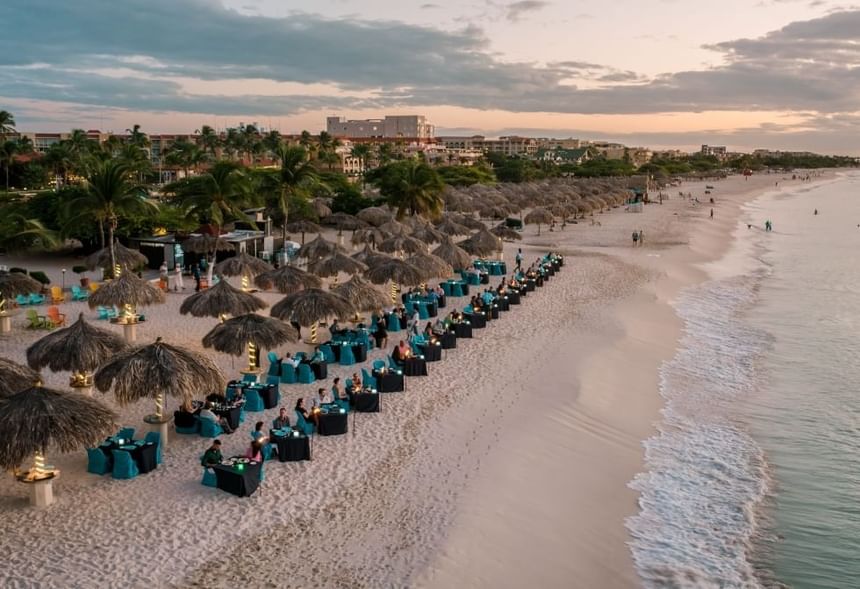 Passions on the Beach  with blue tables and chairs, straw umbrellas, and guests dining on sand by the ocean.
