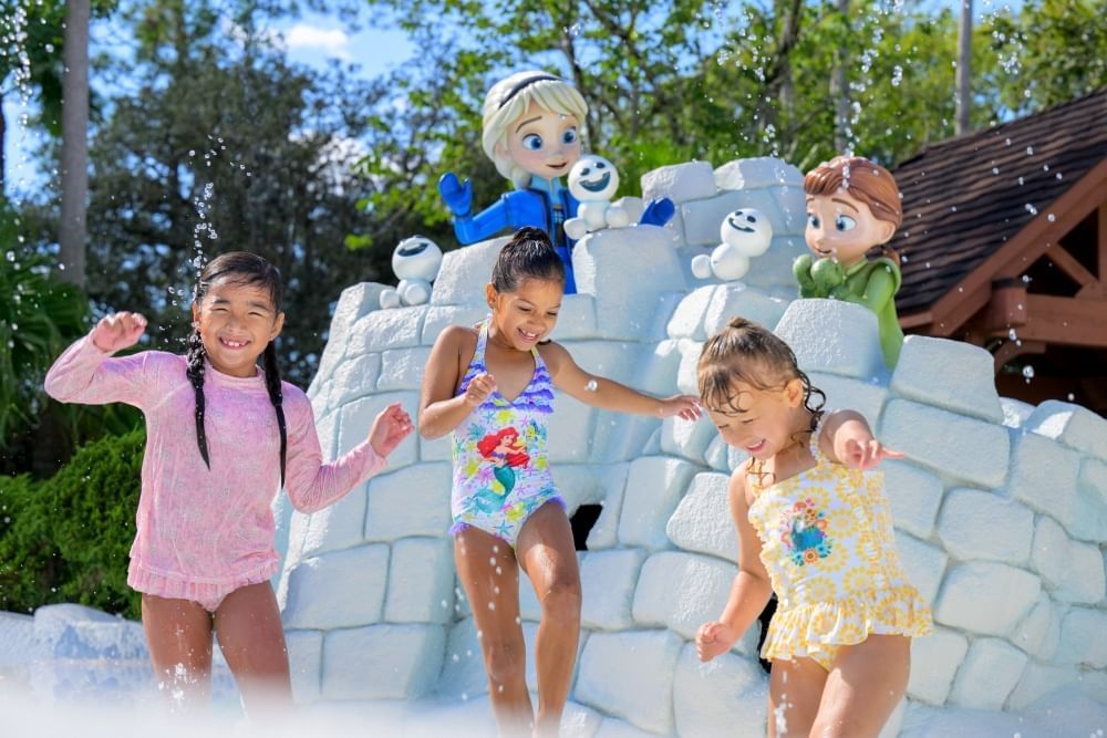 Three girls in bathing suits play in a shallow pool in front of an igloo with statues of young Anna and Elsa from Frozen.