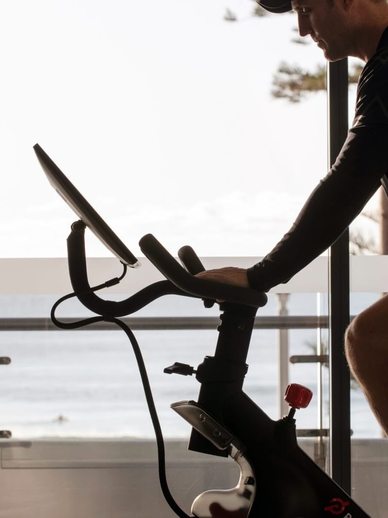 Guest at Elysium Manly riding a stationary bike by a large glass window overlooking a scenic ocean view balcony