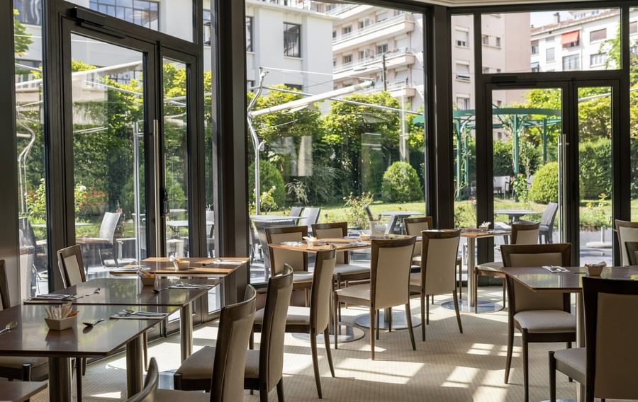 Roseraie Room with wooden tables, beige chairs, & large windows offering a lush garden view at Warwick Reine Astrid - Lyon