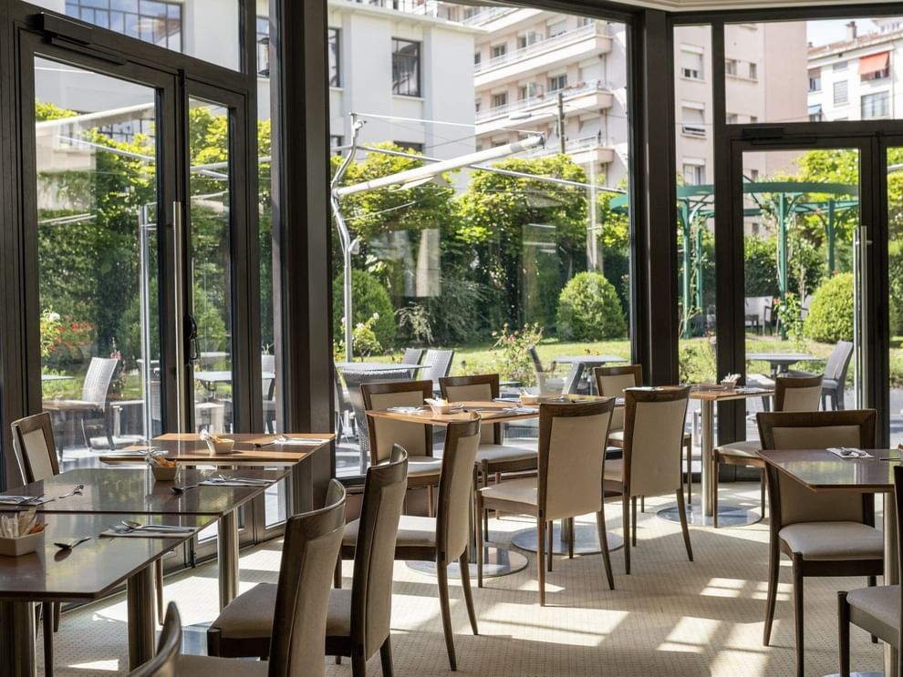 Roseraie Room with wooden tables, beige chairs, & large windows offering a lush garden view at Warwick Reine Astrid - Lyon
