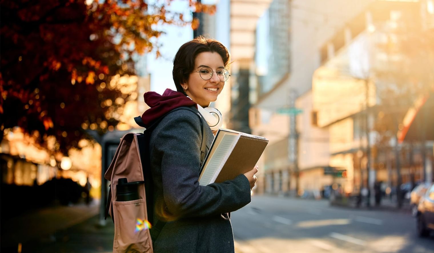 Girl with backpack and headphones holding a book on a sunny city street near UniLodge Canada