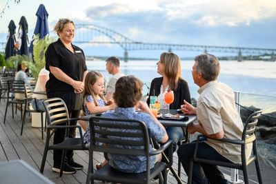 Guests dining on a patio by a steel railing under a cloudy sky, with a bridge surrounding the Rodd Miramichi River