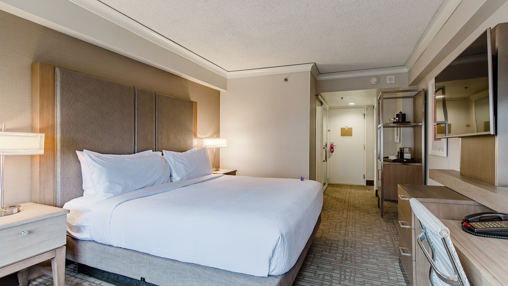 A wide view of a hotel room with a king-sized bed, a large TV mounted on the wall, a desk, and a carpeted floor.