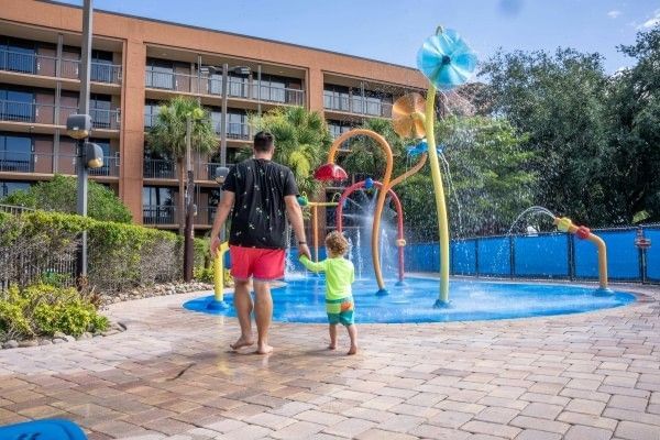 A man holding hands with a toddler walk towards a colorful splash pad at Rosen Inn Lake Buena Vista in Orlando.