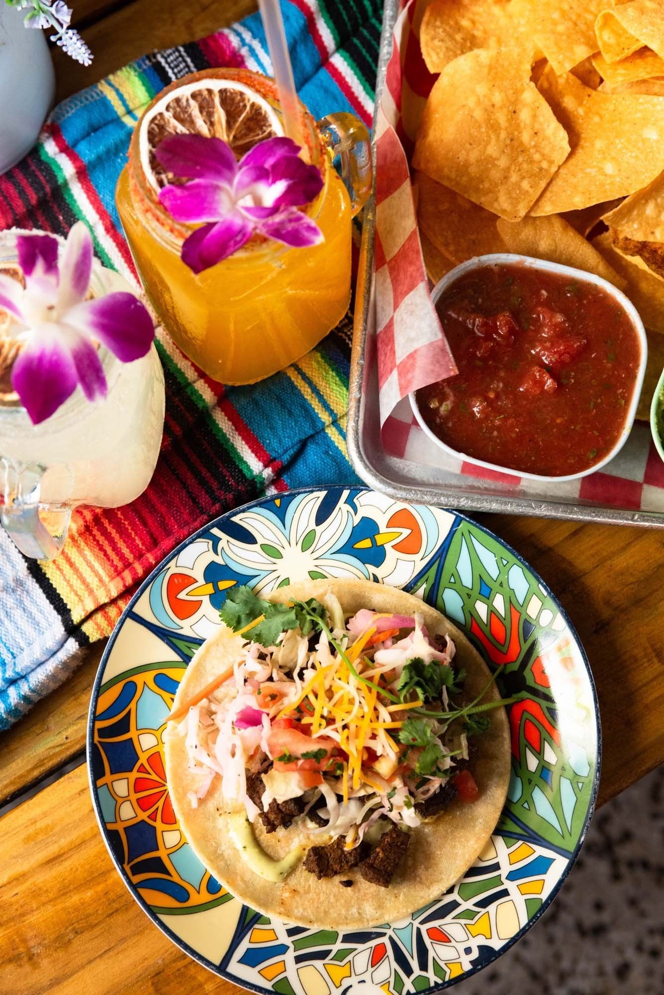 a taco with beef, cheese, and salsa on a colorful plate with a drink and tortilla chips
